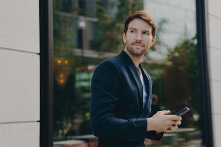 Successful handsome businessman with stubble messaging his employer about project details, holding smartphone in his hands while standing next to building with big window reflecting urban landscapeの写真素材