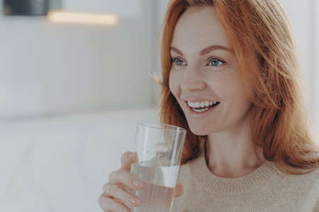 Close up shot of ginger woman drinks pure water from glass enjoys refreshing drink healthy liquid...