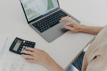 Cropped shot of unrecognizable woman sits at desk with laptop computer calculator and papers...
