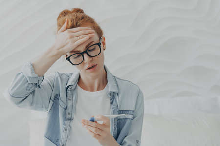 Desperate unhappy young woman sad because of infertility keeps hand on forehead looks at pregnancy test with negative result cannot have baby for long time wears eyeglasses and denim jacket.の写真素材