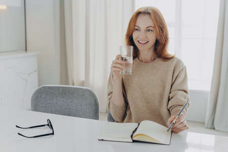 Happy smiling red haired woman housewife dressed casually with glass of pure mineral water enjoying morning at home, writing shopping list in notebook and planning her day, looking aside with smileの写真素材