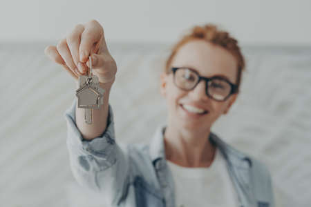 Real estate and relocation concept. Young happy smiling red-haired woman in casual clothes holding house keys of her new house home and showing it at camera, selective focus on female handの写真素材