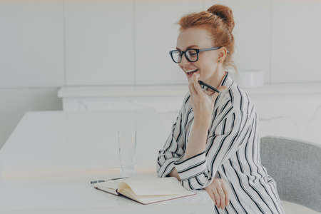 Smiling woman in casual clothes holding mobile and speaking with virtual voice recognition assistant, sitting at her table with notebook and water glass on it, recording audio message on smartphoneの写真素材