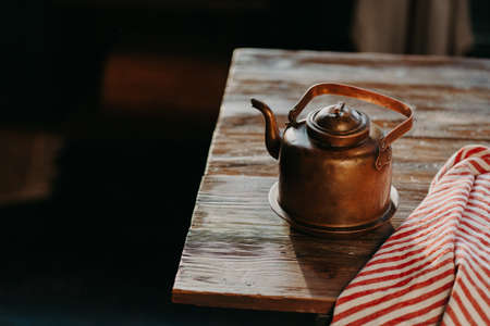 Old copper metal teapot on wooden table in dark room. Red striped towel nearby. Antique kettle for making tea or coffee. Cooking equipmentの写真素材