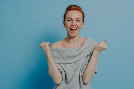 Young overjoyed excited red haired woman dressed in grey knitted sweater clenching fists with...