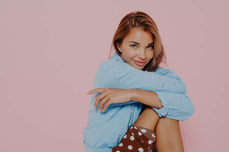 Studio photo of beautiful brunette female sitting and hugging her leg with both hands, slightly...