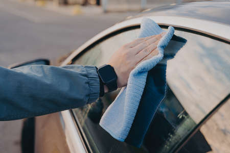 Male hand cleaning car side window with microfiber rag, wiping auto glass dry with cloth during professional vehicle washing outdoors. Car wash conceptの写真素材