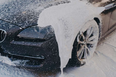Black car in white soap foam at washing station, professional vehicle cleaning with chemical...