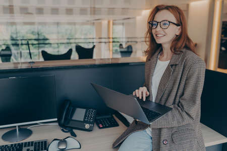 Beautiful woman office worker in casual clothes wearing glasses, sitting on desk at her workplace in...
