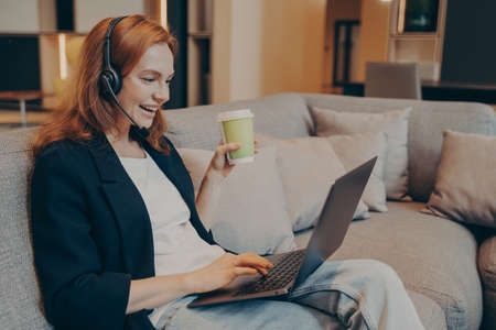 Excited red haired smiling woman with wireless headset and laptop in front of her having online call...