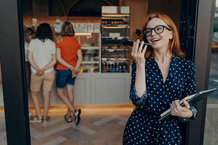 Carefree positive redhead young woman makes voice call keeps smartphone near mouth wears spectacles...