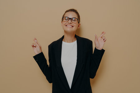 Beautiful happy female keeping her fingers crossed and eyes closed to make desired wish, asking for good luck before important event while posing isolated over studio background. Body language conceptの写真素材
