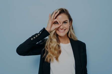 Portrait of beautiful joyful blonde caucasian female wearing black coat looking through okay sign...