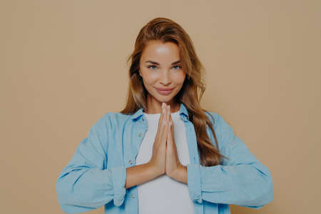 Positive concentrated young woman holding palms together in prayer gesture, being calm and peaceful, making namaste yoga sign on beige background. Body language conceptの写真素材