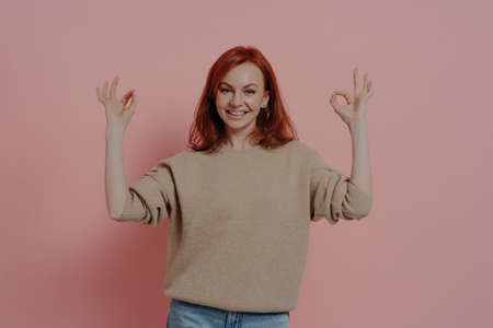 Half length shot of pretty ginger woman showing OK sign with both hands, saying that everything is fine, dressed in casual outfit, satisfied red-haired model posing isolated over pink backgroundの写真素材