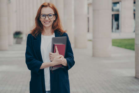 Outdoor portrait of young confident beautiful ginger woman in formal clothes holding laptop and note book and smiling at camera, waiting for business meeting, wearing spectacles, going home after workの写真素材