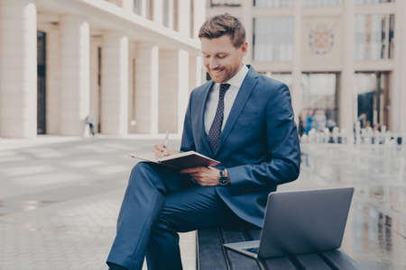 Successful business owner in formal wear writing down information he is receiving from conference call on laptop, sitting on bench with crossed legs and working remotely, doing freelance job outdoorsの写真素材