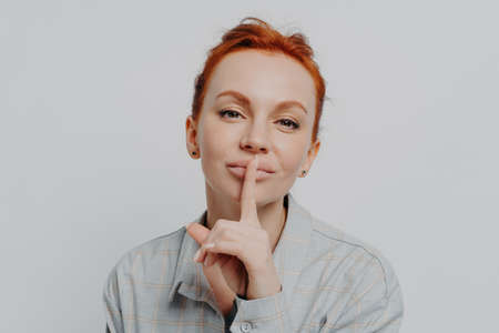 Close up of young redhead woman holding finger on lips, asking to be quiet, keep something in secret, isolated on grey studio background, caucasian female showing shh shush gestureの写真素材