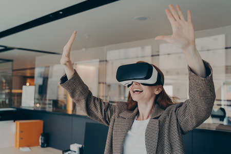 Young excited redhead businesswoman in formal wear sitting in office interior using VR headset and...