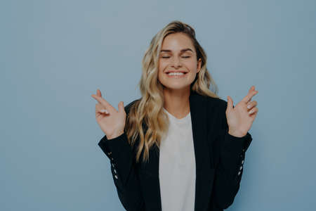Superstitious young student girl with blond hair crossing fingers with both hands, invoking good luck before important exam or event, hopes for victory and success, feeling hopeful closing her eyesの写真素材