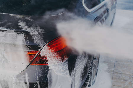 Carwash equipment. Vehicle covered in white soapy foam during regular wash outdoors with cleaning products and chemicals, spraying detergent with high-pressure washer on car exterior surfaceの写真素材