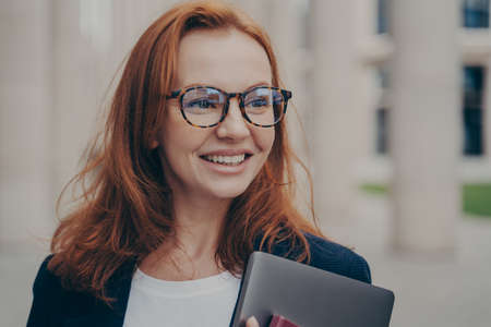 Portrait of happy elegant female with broad smile looking aside while waiting for business partner outdoors, holding modern laptop and wearing spectacles, ginger business woman going home after workの写真素材