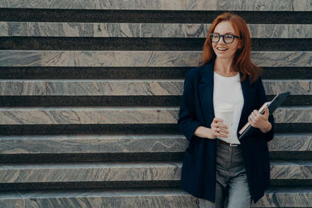 Horizontal shot of cheerful positive young redhead woman dressed formally drinks takeaway coffee...