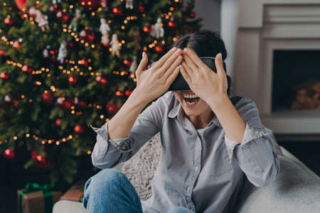 Joyful young woman in VR headset glasses sitting on cozy sofa next to decorated Xmas tree, amazed happy female in virtual reality googles closing eyes with hands, enjoying christmas holidays at homeの写真素材