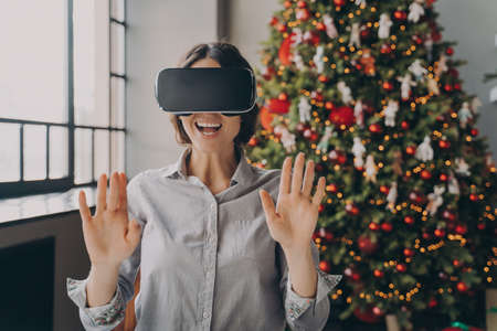 Impressed europian lady using VR googles while spending Christmas at home, playing 3D games in room with beautiful lush Xmas tree on background, happy woman completely immersed in virtual worldの写真素材
