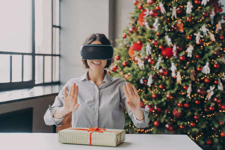 Joyful hispanic female in VR headset sitting at desk with Xmas present during Christmas holidays,...