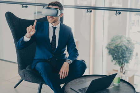 Smiling bearded businessman using modern VR headset glasses while sitting in office lobby and...