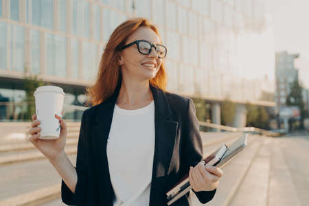 Overjoyed happy ginger woman in spectacles drinking coffee and holding laptop while walking outdoors...