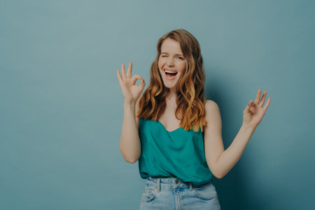 Charming funny girl showing OK sign with both hands while winking and looking at camera with big...