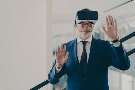Young male employee in formal suit using vr technologies at work, businessman in virtual reality glasses standing on stairs in office, trying to touch objects in digital actuality with both handsの写真素材