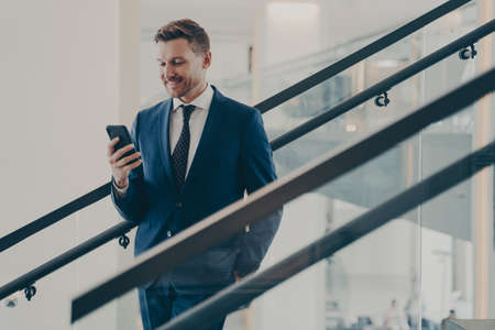 Happy bearded businessman with smartphone standing on staircase. Satisfied young man using mobile phone app, browsing internet, looks at smartphone with smile. Mobile technology and business conceptの写真素材