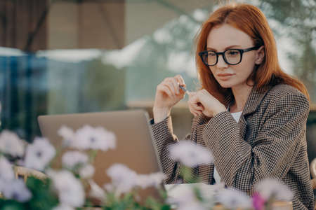 Young focused red-haired business woman works online at outdoor cafe, female in eyeglasses looking at laptop screen with pen in hands during remote work or distant learning at coffee shopの写真素材
