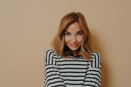Feeling shy. Young charming woman with dyed blonde hair in striped shirt playfully smiling while looking at camera with head slightly down, isolated over beige studio background with free spaceの写真素材