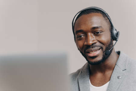 Young pleased african american man office worker in headset communicating with colleagues during video call on laptop, smiling guy call center agent talking with client, consulting customer onlineの写真素材