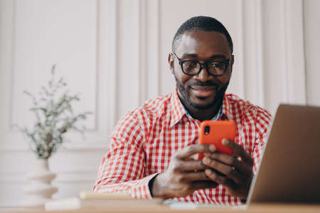Cheerful smiling African ethnicity man office employee sitting at desk with open laptop holding smartphone chatting with friends while working remotely online from home. Freelance conceptの写真素材