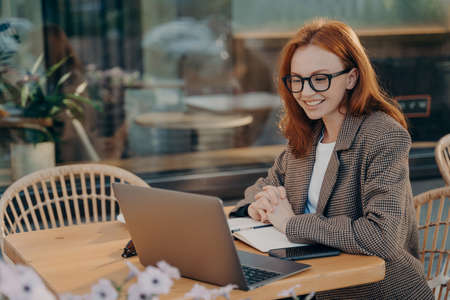 Redhead female freelancer watches training webinar makes notes in notepad sits in front of opened laptop computer dressed formally poses at outdoor cafe browses internet develops new projectの写真素材