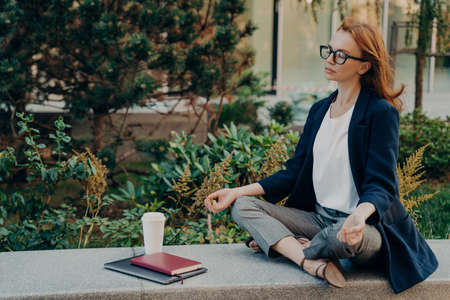 No stress. Calm red-haired woman with closed eyes relaxing while working remotely on laptop outdoors, mindful young business woman meditating with closed eyes, doing breathing yoga exercises outsideの写真素材