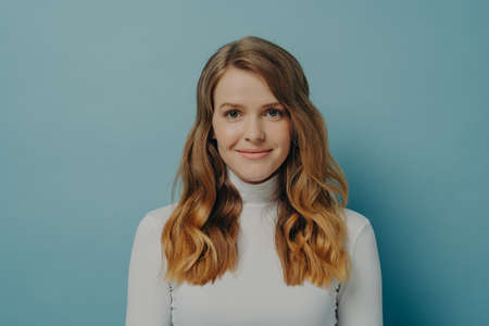 Half-length portrait of beautiful dreamy millennial woman with wavy hair in white turtleneck looking at camera with gentle smile isolated over pastel blue studio wall background with copy spaceの写真素材