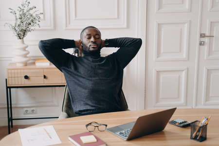 Tired overworked african american male office worker with eyes closed and hands behind head relaxing at workplace, male freelance employee sitting at desk with laptop and resting after work doneの写真素材