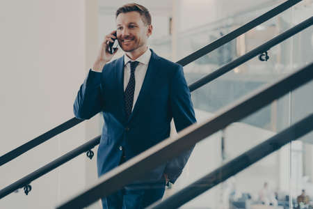 Handsome smiling european businessman in elegant blue suit having mobile conversation with client, making pleasant business call while standing on stairs in office, talking on smartphone at workの写真素材