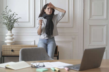 Worried unhealthy freelancer lady checks thermometer wearing warm knitted scarf feeling hot from...