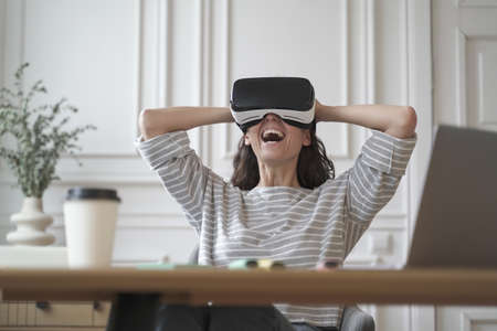 Amazed woman office employee in VR glasses laughing with joy leaning back in chair with hands behind head while remotely working from home office, cheerful lady spends free time in augmented realityの写真素材