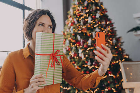 Joyful Spanish bright brunette lady making selfie on smartphone while posing with Xmas gift close to...