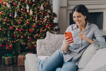 Happy female in wireless earbuds holds smartphone in hands, talking with family or friends online at Christmas time, sitting on cozy sofa in living room at home with bright colored Xmas treeの写真素材