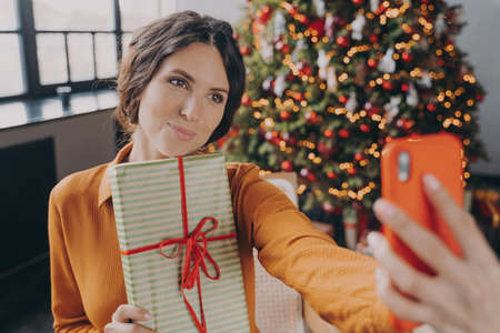Happy charming young brunette woman taking selfie with Christmas gift box on smartphone while...