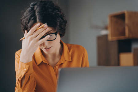 Frustrated tired young Spanish woman office worker in glasses covering face with hand while sitting...